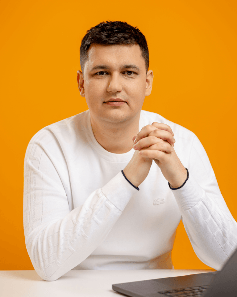 Чоловік у білому светрі сидить за столом зі зчепленими руками, обличчям до камери. У нього коротке темне волосся і нейтральний вираз обличчя. На столі стоїть ноутбук, фон - яскраво-помаранчевий.